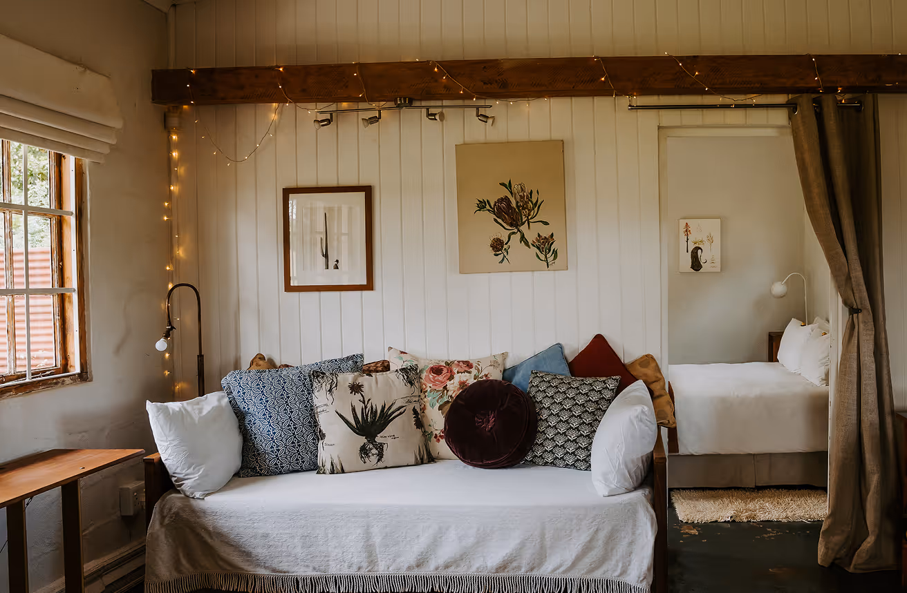 Cozy living room with a white-covered wooden sofa adorned with assorted patterned cushions, a wooden side table, fairy lights, and a glimpse into a bedroom with a white bed and wall art.
