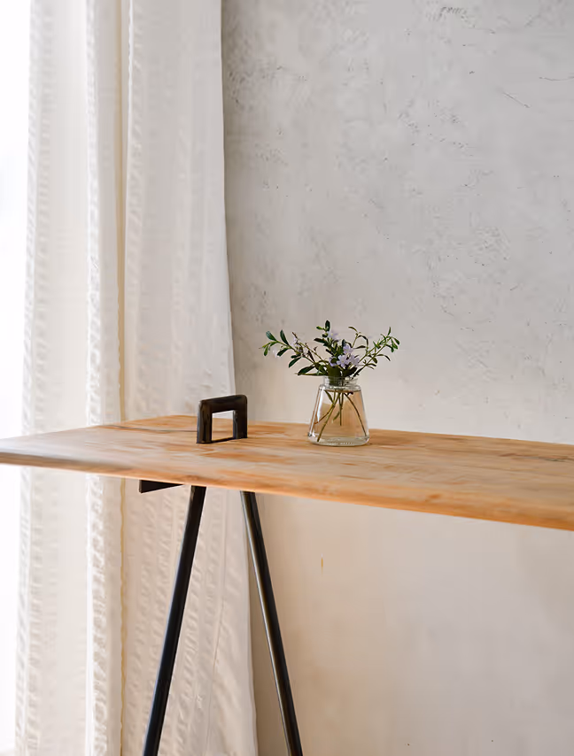 Minimalist wooden desk with black metal legs and a small glass vase holding green branches with tiny purple flowers, set against a textured light wall and beige curtain.