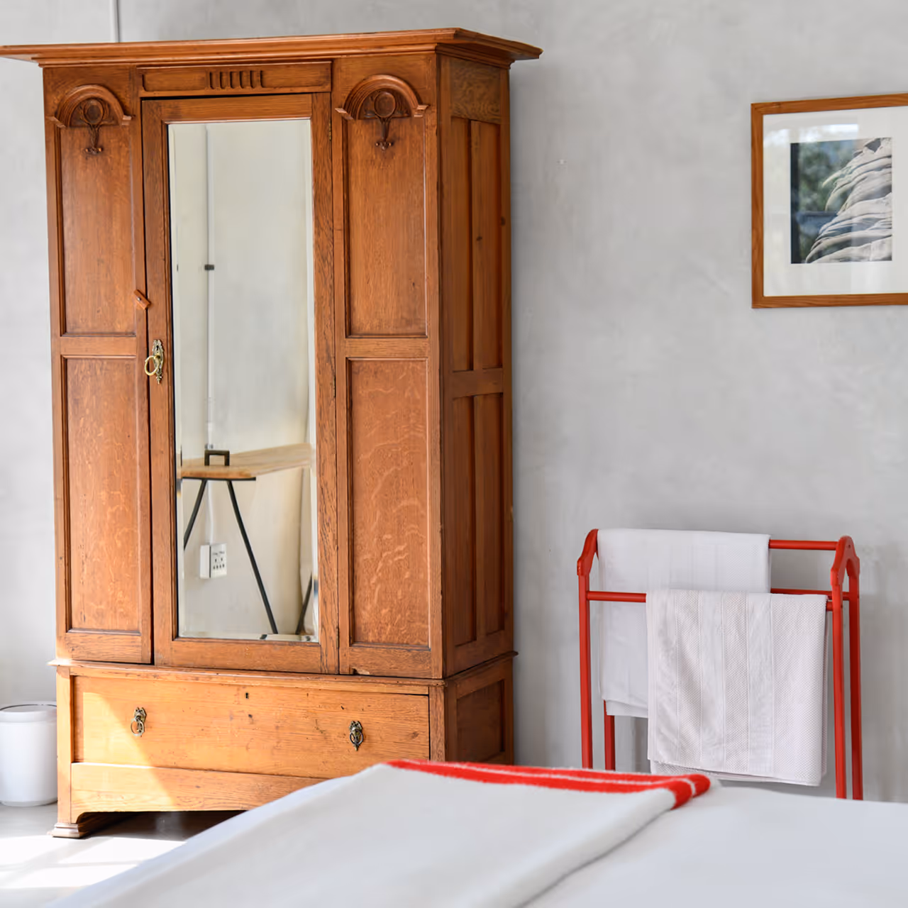 Wooden wardrobe with a central mirror in a minimalistic room with a red towel rack holding white towels and a framed picture on the wall.