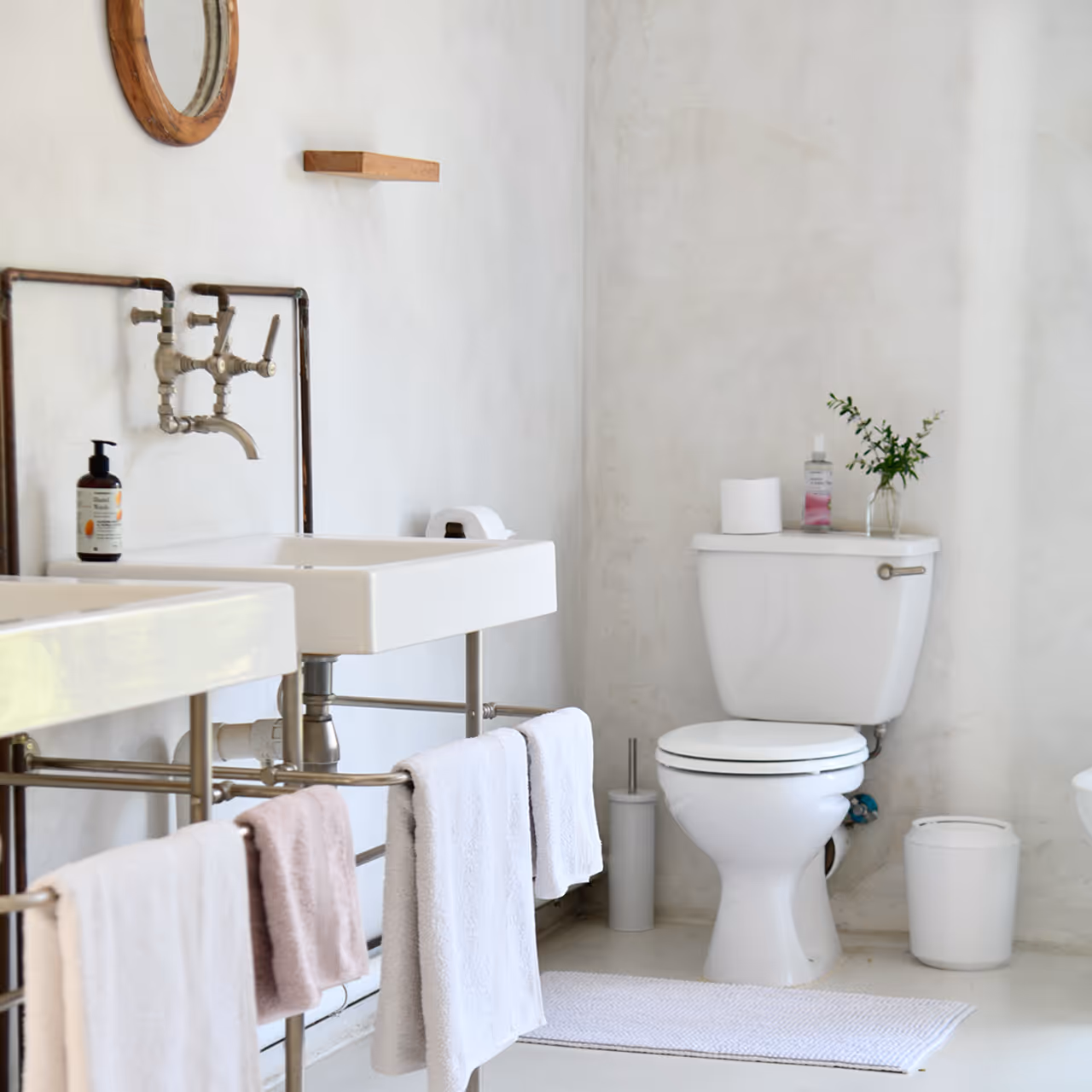 Minimalist bathroom with white toilet, two sinks with towel racks and metal faucets, and a small round mirror on the wall.