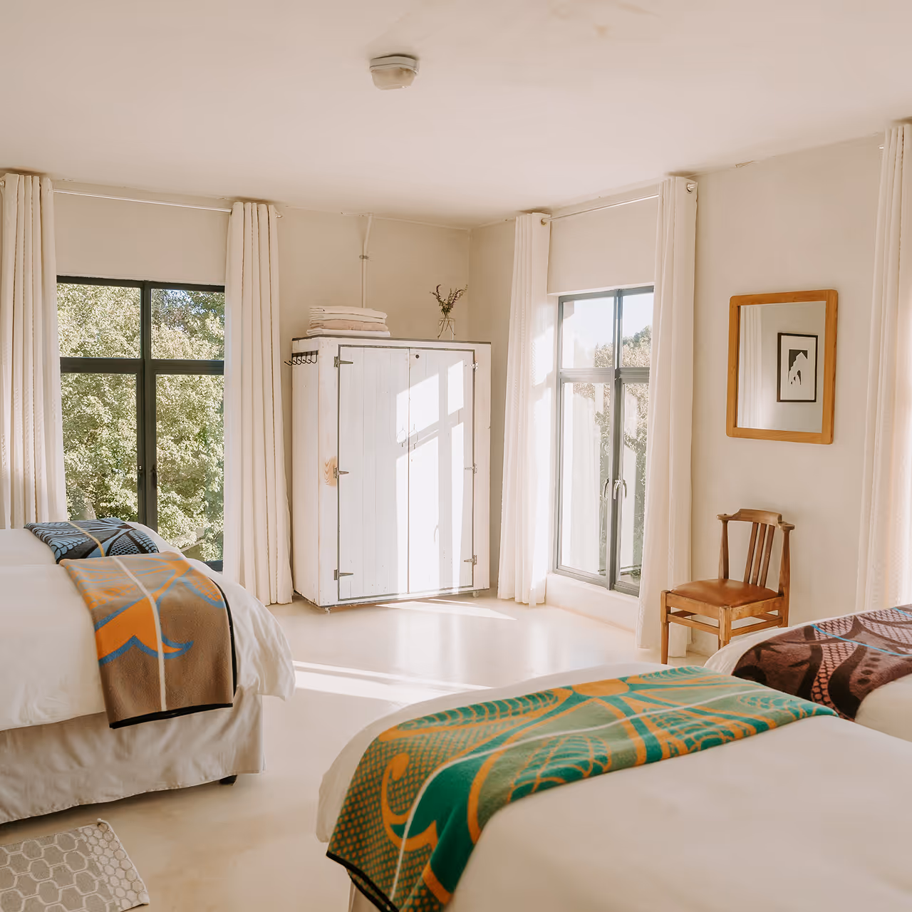 Bright bedroom with two beds each covered with colorful blankets, large windows with white curtains, a white wardrobe, and a wooden chair under a framed mirror.