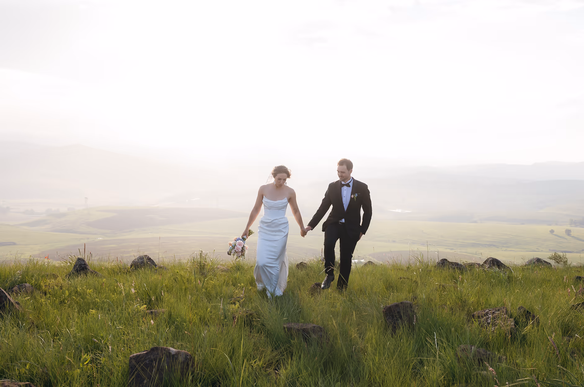 Bride and groom holding hands and walking through a green field with rocks under a bright sky.