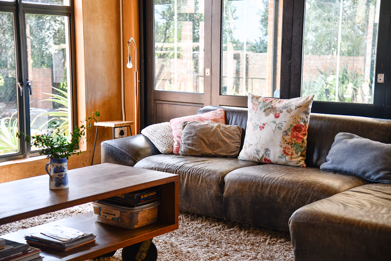 Cozy living room with a dark brown leather sectional sofa adorned with various cushions, a wooden coffee table with a blue and white vase holding green plants, and large windows letting in natural light.