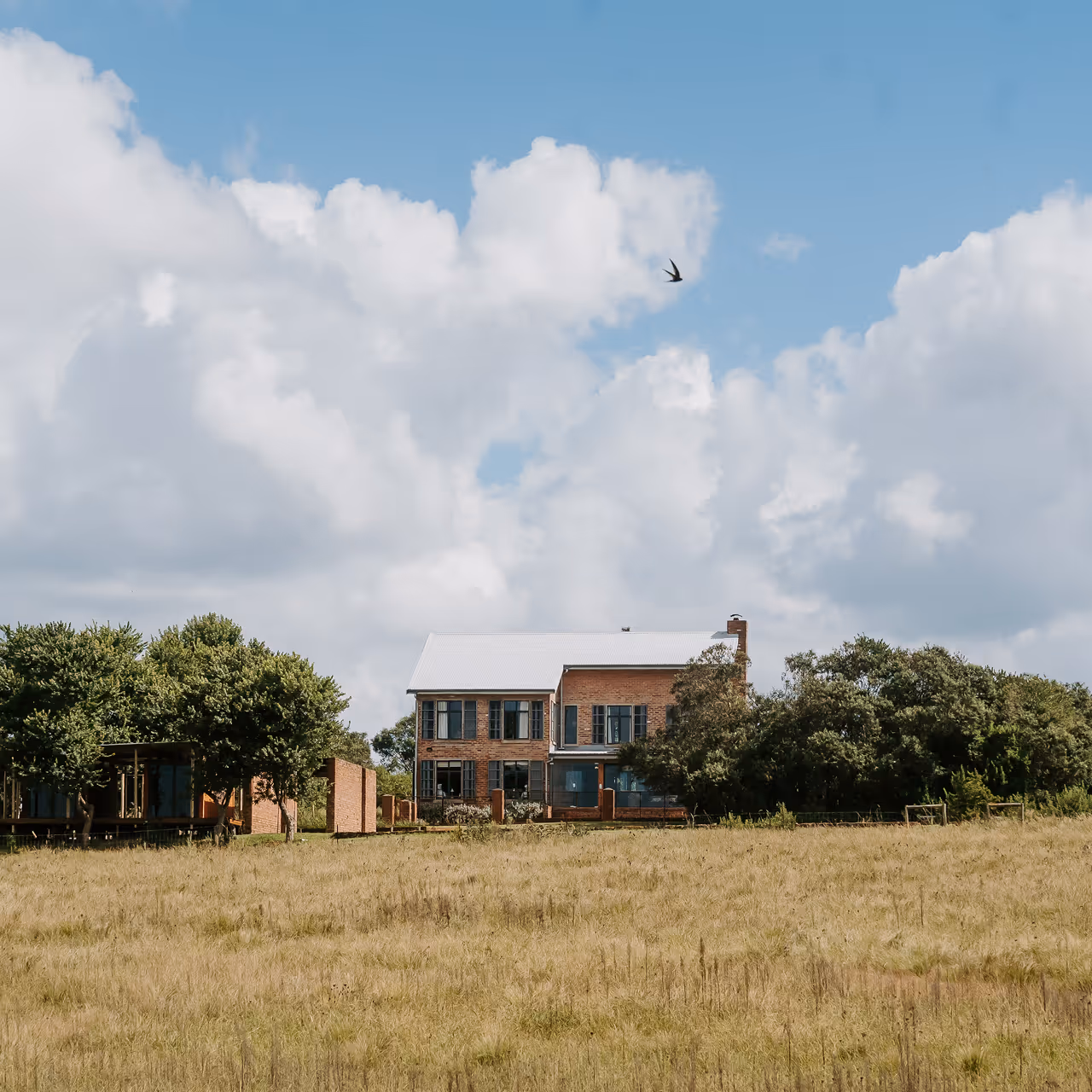 Brick house with a white roof surrounded by trees behind a grassy field under a partly cloudy sky with a bird flying.