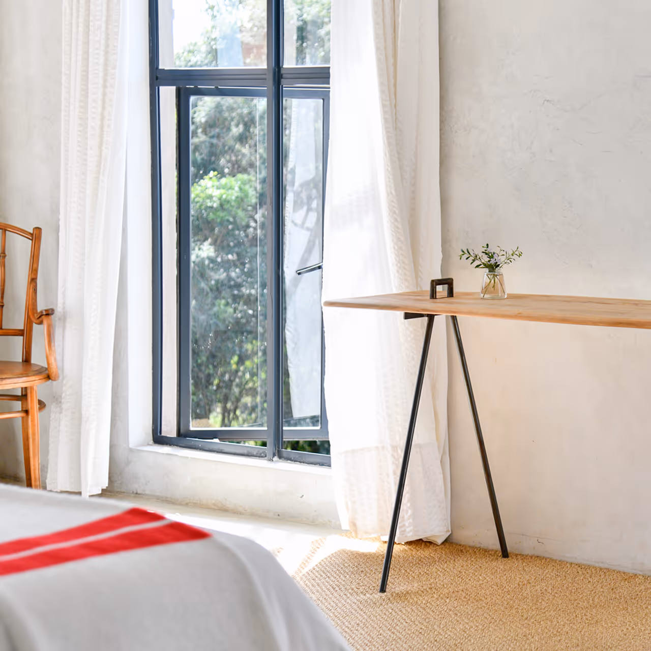 Bright room corner with an open black-framed window, wooden chair, simple wooden table with a small vase of greenery, and a white curtain.