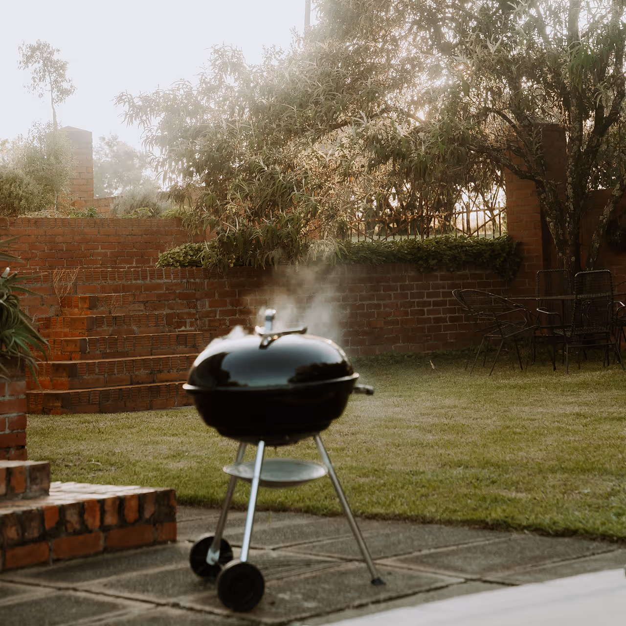 Black charcoal grill emitting smoke on a patio with a green lawn and brick walls in the background.