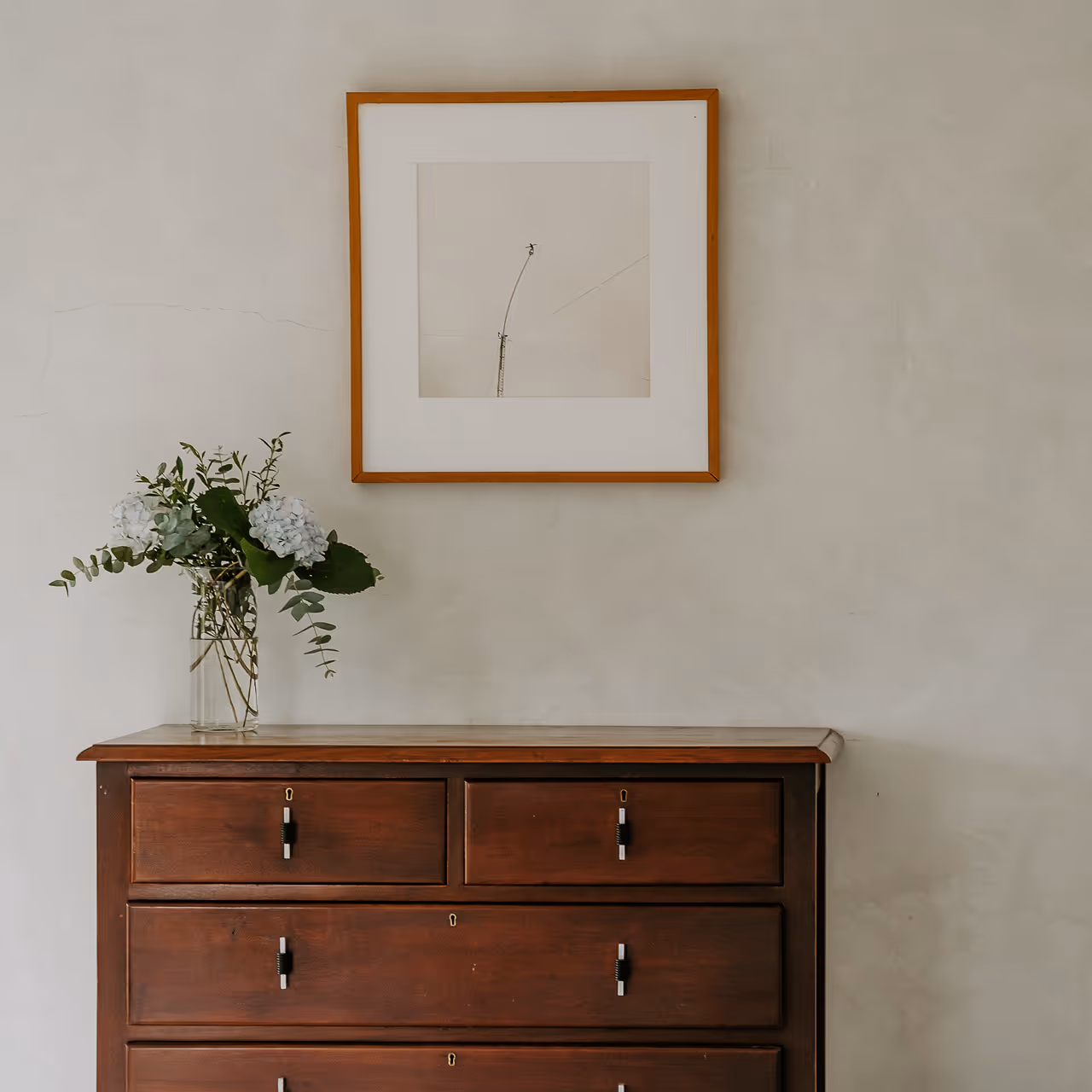 Wooden dresser with four drawers topped by a vase of white flowers and greenery, beneath a framed minimalist artwork on a beige wall.