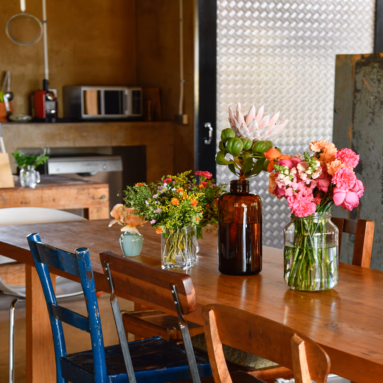 Wooden dining table with various colorful flower arrangements in glass vases and mismatched chairs in a rustic kitchen setting.