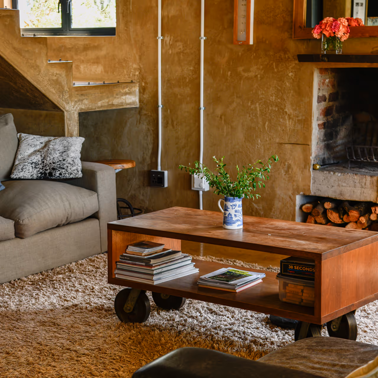Cozy living room with gray sofa, wooden coffee table with books and green plant, shaggy rug, and fireplace with stacked firewood.