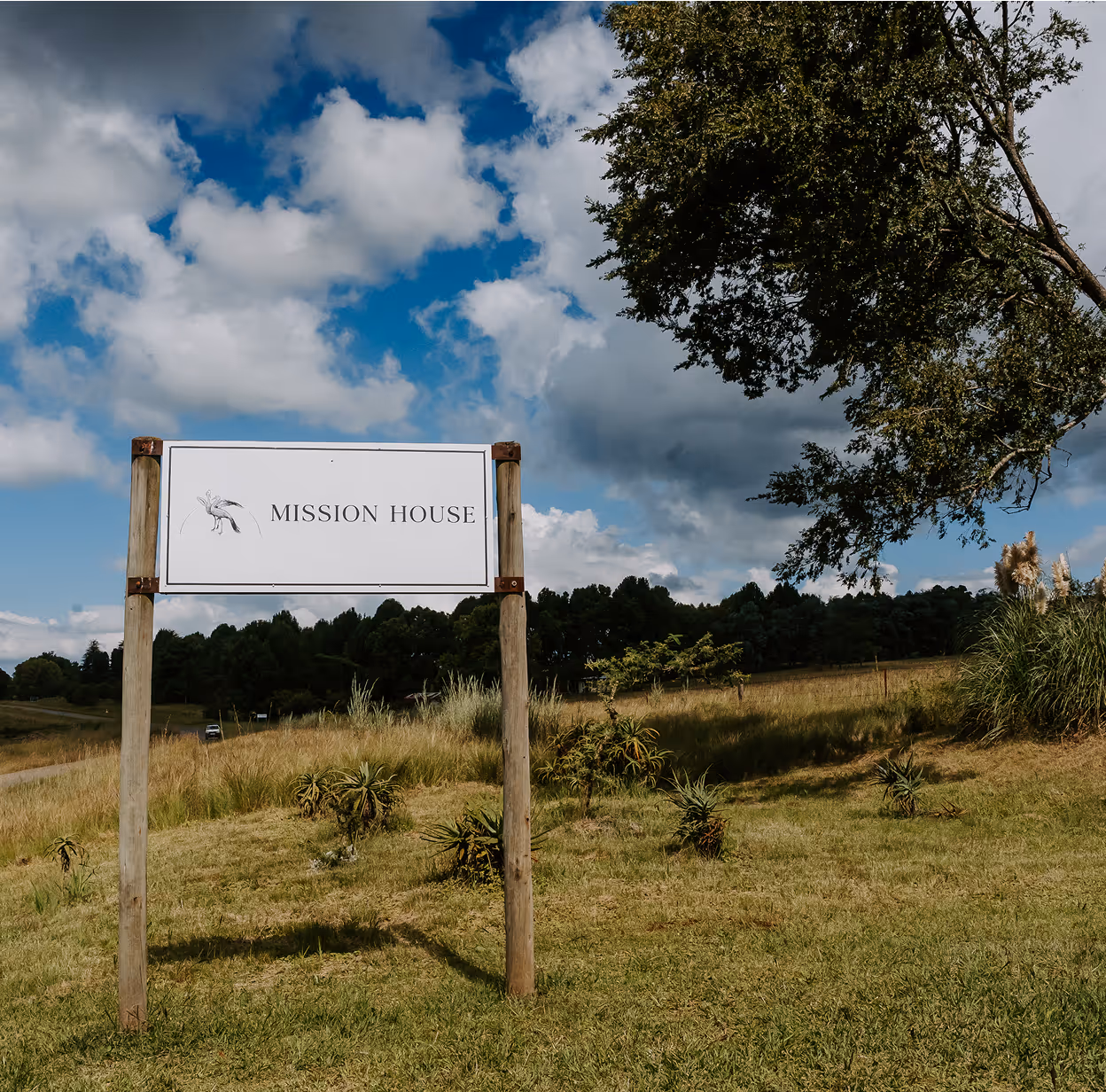 White rectangular sign on wooden posts in a grassy field with trees and a cloudy blue sky, reading 'MISSION HOUSE'.