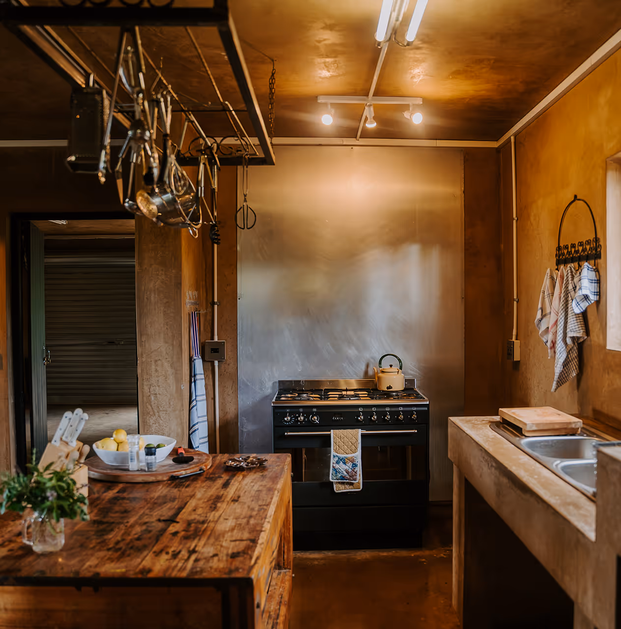 Rustic kitchen with wooden countertops, a black stove with a beige kettle, hanging utensils, and dish towels on the wall.