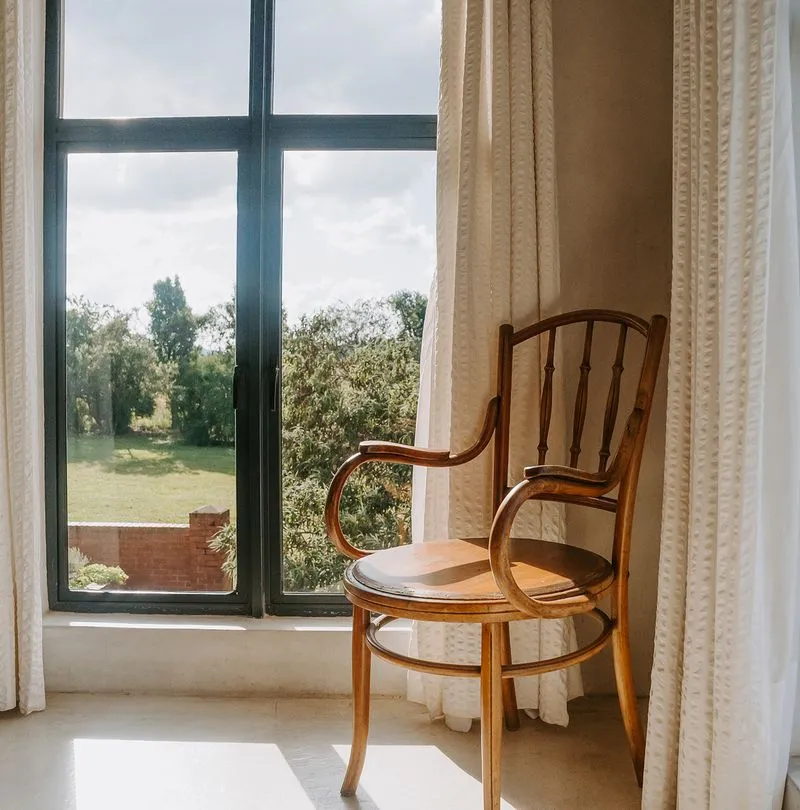 Wooden chair with curved armrests in front of a large window with white curtains and a view of green trees and grass outside.