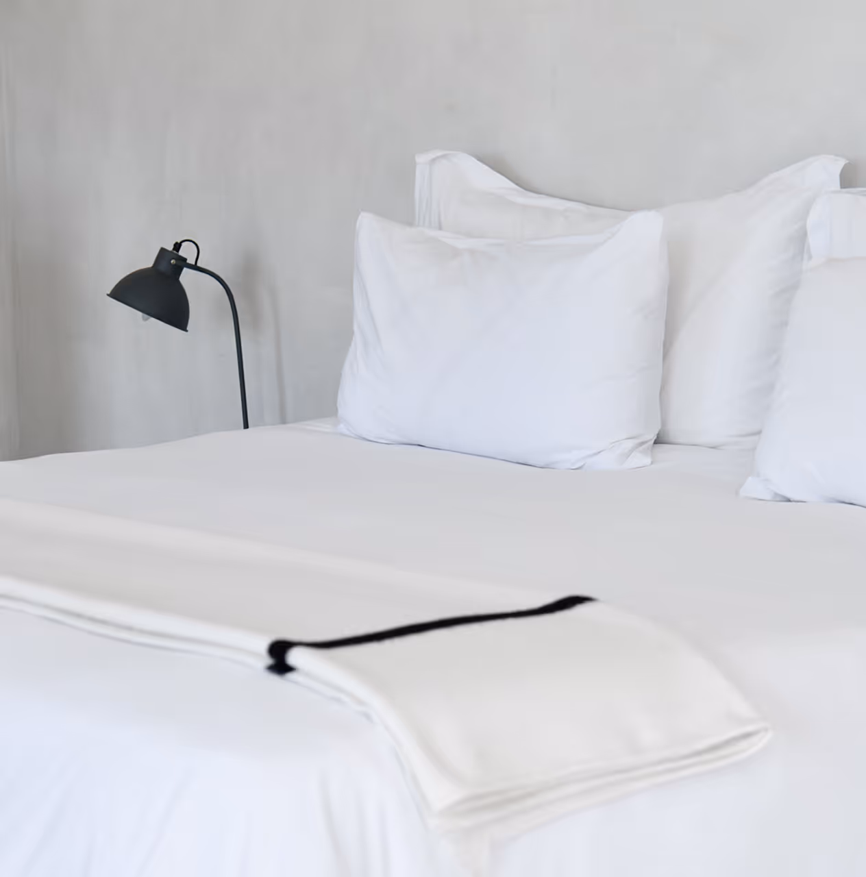 Neatly made bed with white pillows, white linens, and a folded white blanket with a black stripe, next to a black bedside lamp.