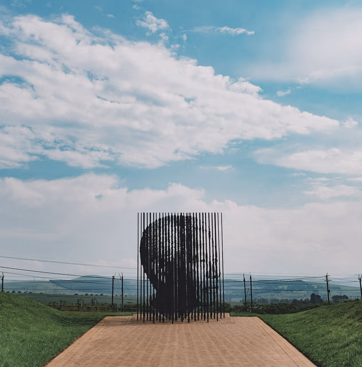Sculpture of a face formed by spaced vertical black rods on a paved path with grassy sides under a partly cloudy blue sky.